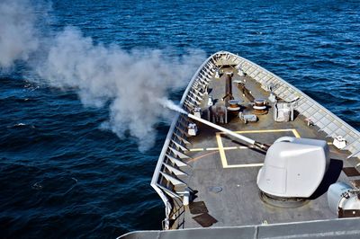 Aerial view of a naval vessel firing, with a plume rising over blue water